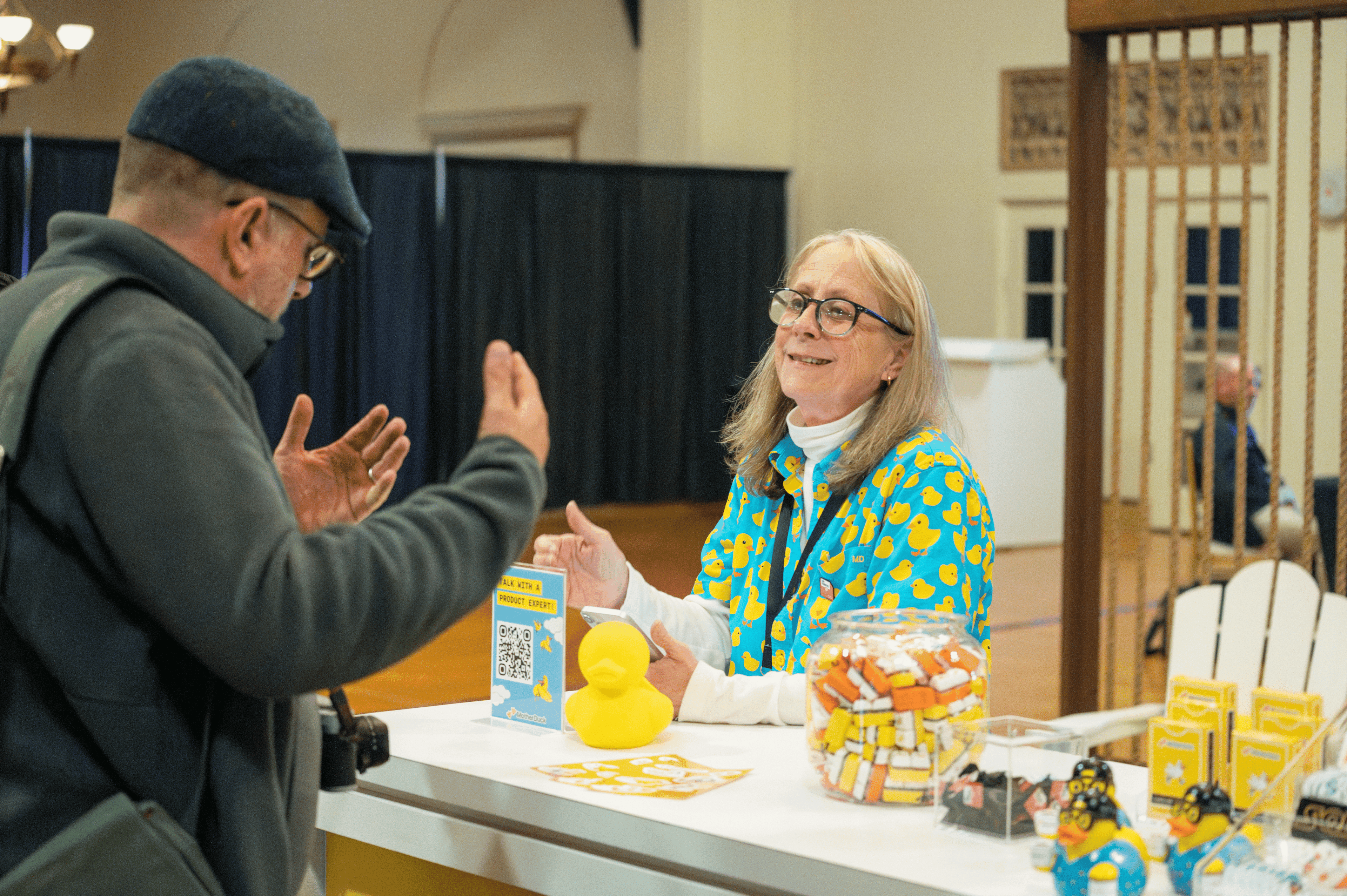Woman in duck-patterned shirt converses with man at a table with rubber ducks.
